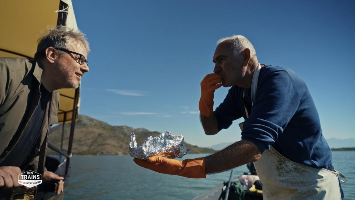 Philippe Gougler face à un pêcheur lui proposant de goûter de la truite cuite